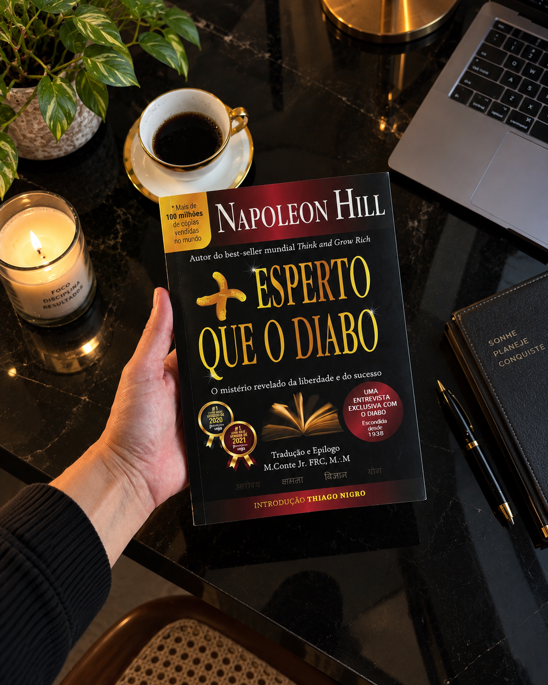 Livro do mês Mais Esperto que o Diabo, de Napoleon Hill, segurado sobre uma mesa com café, vela e notebook